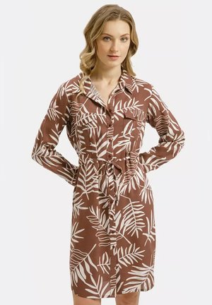 Kobieta ubrana w brązową sukienkę na guziki z białym wzorem liści i przewiązaną w talii, stojąca z rękami na biodrach na tle jednolitego tła.