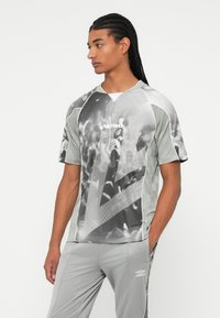 Fiatal férfi plettyes hajjal, szürke Umbro sportpólóban, amely absztrakt tömeg mintázattal rendelkezik, és hozzá illő szürke Umbro nadrágban áll, keze a zsebében.