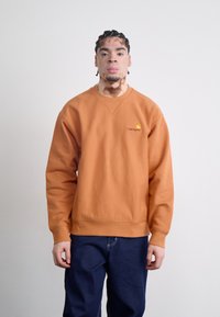 Orange sweatshirts med ribbede ærmer og kant, der har et lille gult logo på venstre bryst. Kombination med blå denimbukser.