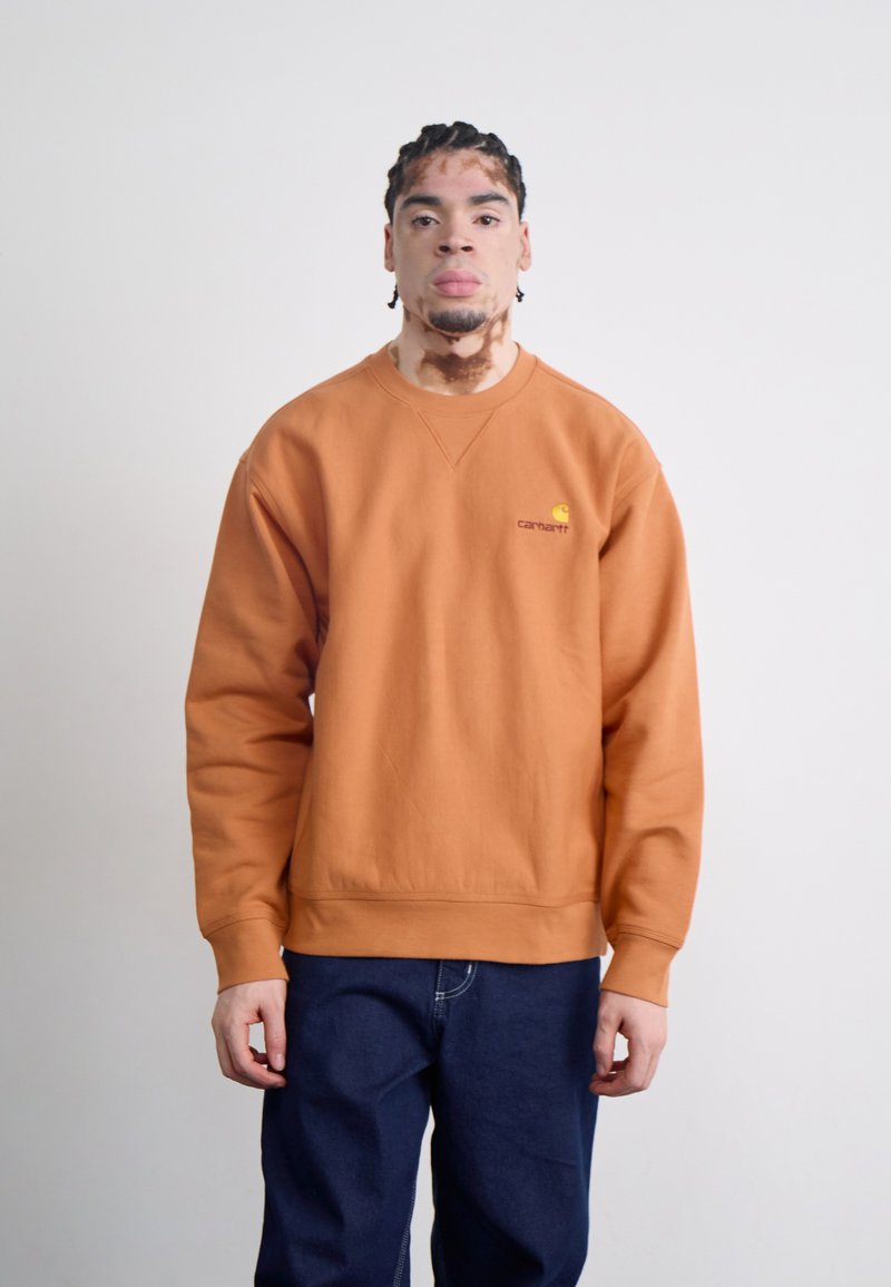 Orange sweatshirts med ribbede ærmer og kant, der har et lille gult logo på venstre bryst. Kombination med blå denimbukser.