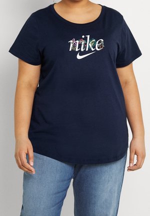 Osoba nosząca granatowy t-shirt Nike z kwiatowymi wzorami na logo, zestawiony z jasnoniebieskimi jeansami, stojąca na jednolitym tle.