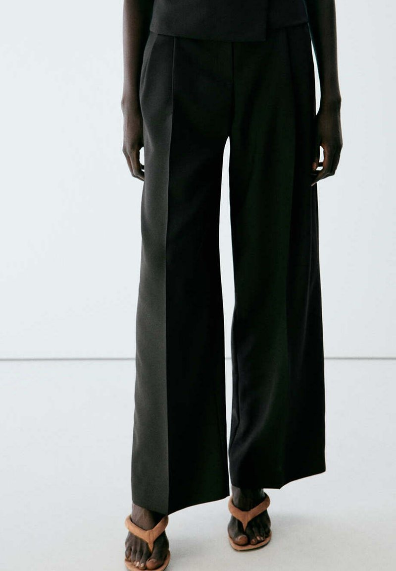 Personne portant un pantalon noir à jambes larges et des sandales beige ouvertes à l'avant, debout sur un fond clair uni.