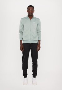 Jachetă bomber verde deschis cu fermoar, manșete cu nervuri și logo EA7, asortată cu pantaloni de trening negri și adidași albi pe un fundal alb.