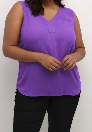 Kobieta ubrana w fioletowy top bez rękawów z dekoltem w serek i czarne spodnie, delikatnie splecione dłonie na talii na jednolitym tle.