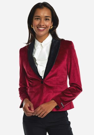 Red velvet blazer with black shawl lapels, single button closure, and two front pockets. Worn over a white ruffled shirt.