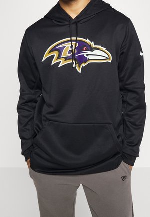 Man wearing black hoodie with a large purple and gold Baltimore Ravens logo on the chest and gray pants with a small logo.