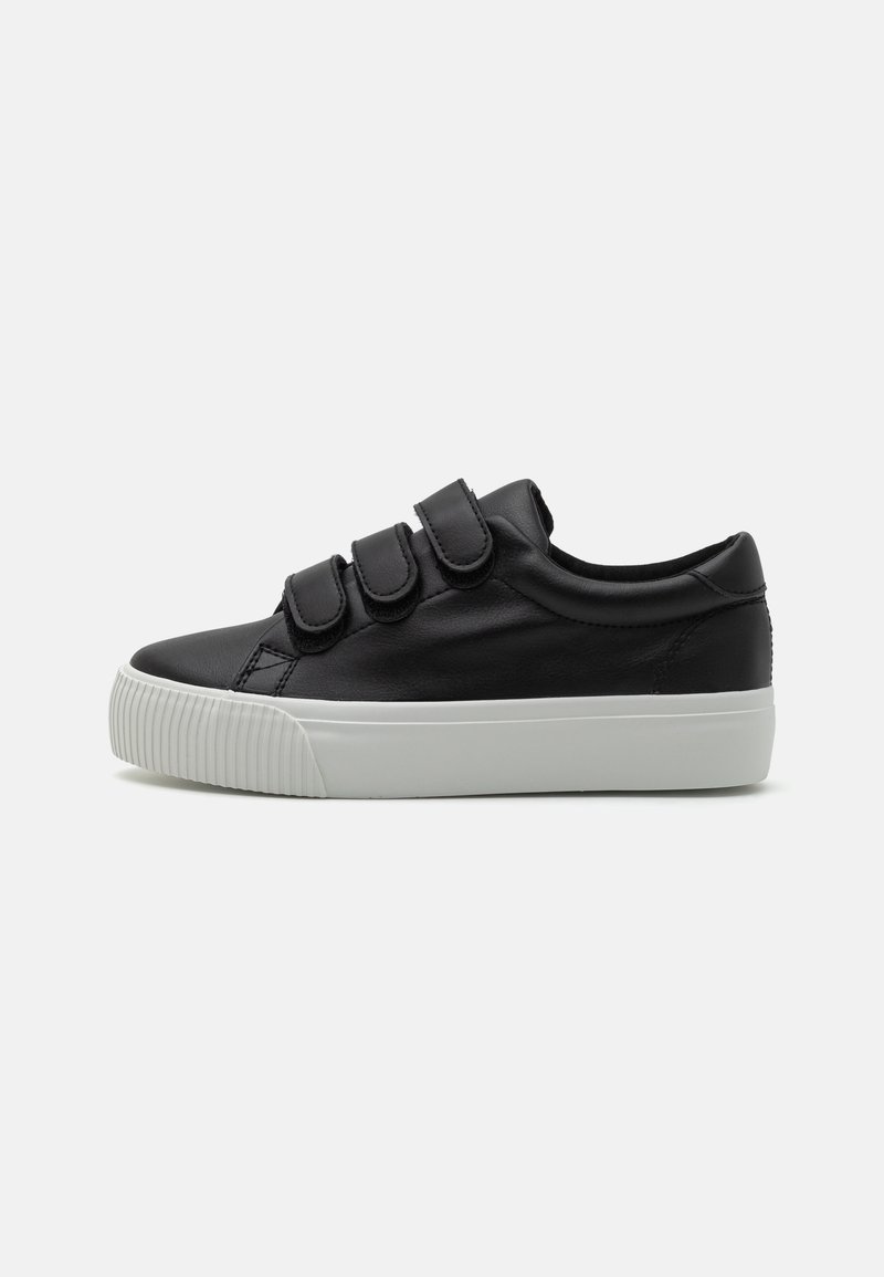 Zapatilla negra de cuero con tres cintas de Velcro ajustables, suela de goma blanca y textura estriada a lo largo del borde de la suela.