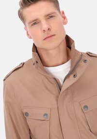 Tan lynlåsjakke med høj krave, to forlommer, sølvfarvede detaljer og en tekstureret vævning. Båret over en hvid skjorte.