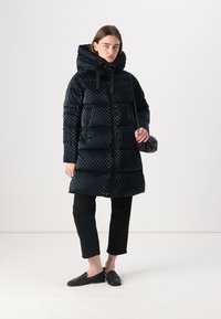 Cămașă puffer de culoare albastră închisă, cu un model în carouri, având un guler înalt și glugă, fermoar negru și două buzunare laterale. Pantaloni și pantofi negri.