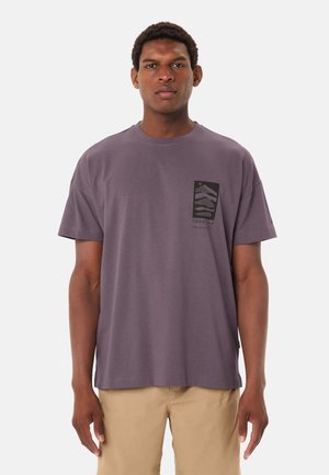 Uomo che indossa una T-shirt color lavanda con stampa grafica nera astratta e pantaloni beige, in piedi davanti a uno sfondo bianco semplice.