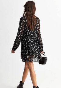 Black floral dress made of sheer fabric, featuring long sleeves, a flared hem, and white flower patterns. Accessory: small black handbag.