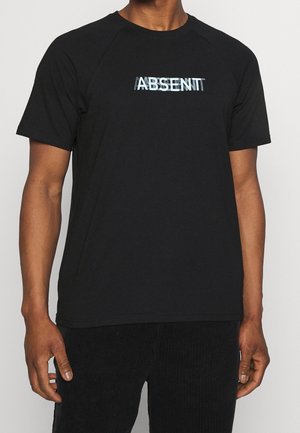 Homme portant un t-shirt noir avec un texte blanc déformé "ABSENT" sur la poitrine, associé à un pantalon noir.