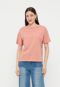 Γυναίκα με μακριά σκουρόχρωμα μαλλιά φοράει κοραλί κοντομάνικη μπλούζα και μπλε τζιν, στέκεται μπροστά σε ένα απλό φωτεινό φόντο, κοιτάζοντας μπροστά.