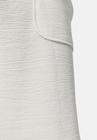 Tejido blanco texturizado con un patrón acanalado, que presenta un detalle de bolsillo doblado de un lado. El material parece suave y estructurado.
