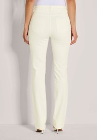 Vrouw draagt hoge taille, recht gesneden crèmekleurige broek met achterzakken en witte hoge hakken, gezien van achteren.