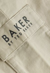 Beige kangasta, jossa on hienovarainen pinta, ja siinä on suorakulmainen tarra, jossa lukee "BAKER by TED BAKER" paksuilla mustilla kirjaimilla.