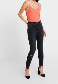 Jeans negros de tiro alto con un ajuste ceñido, con un pequeño desgarrón en la rodilla. Combinados con una camiseta sin mangas naranja y zapatos de tacón alto negros.