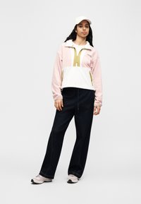 Γυναίκα που φοράει ροζ και λευκό μπουφάν με φερμουάρ, μαύρο παντελόνι με φαρδιά μπατζάκια, ανοιχτό ροζ αθλητικά παπούτσια και λευκό καπέλο, στέκεται μπροστά σε απλό φόντο.