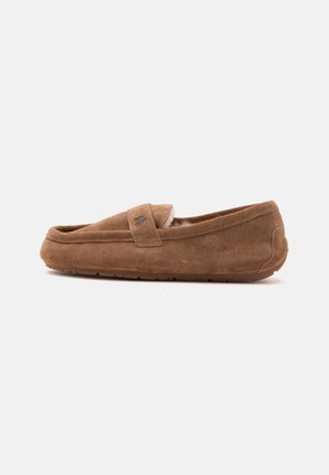 Chausson mocassin en daim marron avec doublure en fourrure douce et semelle en caoutchouc, vu de côté sur un fond blanc uni.