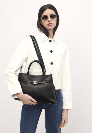 Black leather handbag with dual handles, silver hardware, and a structured shape. Model wears a white jacket and sunglasses, posing against a neutral background.