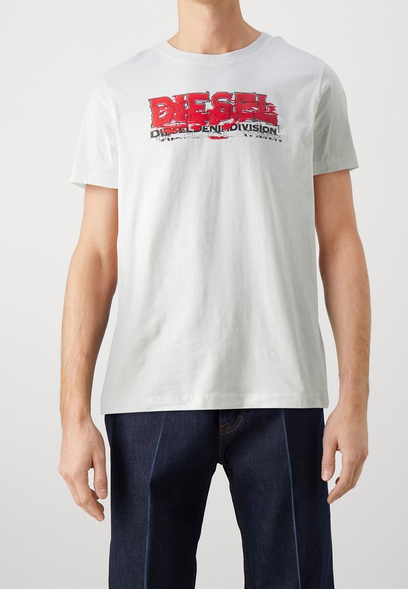Hombre con camiseta blanca con gráfico de texto rojo y negro "DIESEL" y jeans azul oscuro, de pie frente a un fondo blanco liso.