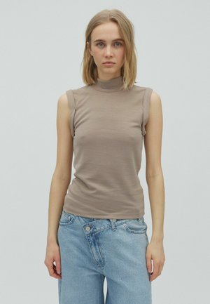 Tânără cu păr blond poartă un top bej fără mâneci și blugi lejeri, de culoare albastru deschis, pe un fundal gri simplu.