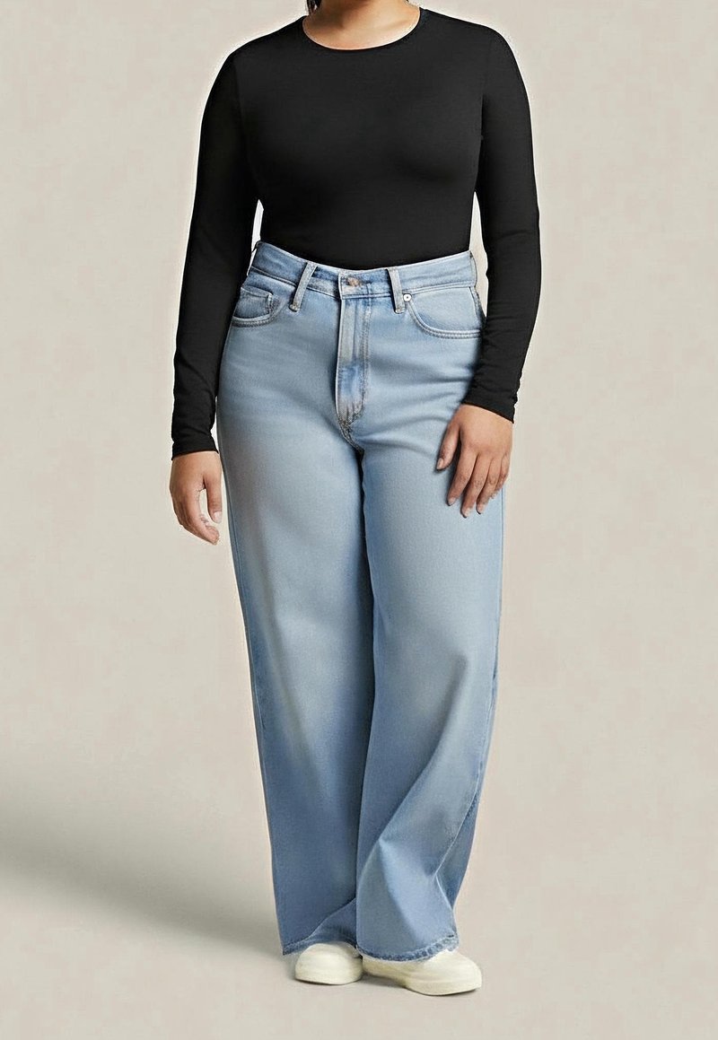 Personne portant un haut noir à manches longues, un jean bleu clair taille haute à jambes larges et des chaussures blanches, debout devant un fond beige uni.