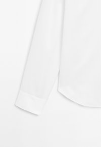 Camicia bianca a maniche lunghe con texture liscia, orlo arrotondato e dettagli dei polsini abbottonati, caratterizzata da un design classico e sartoriale.