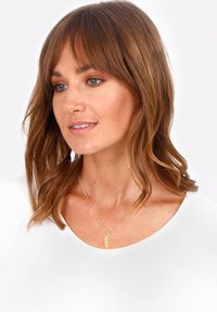 Gouden veerhangersketting met een kleine juweelaccent op een delicate ketting, gedragen over een wit topje, met zacht, golvend haar.