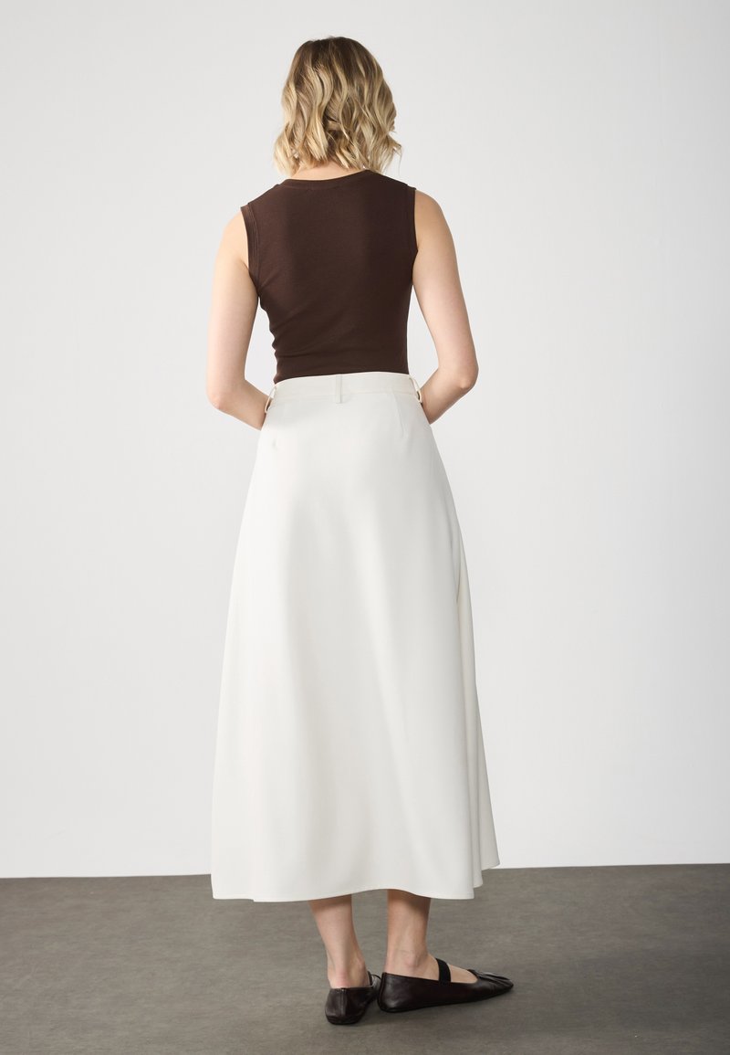Ärmelloses braunes Top kombiniert mit einem fließenden weißen Midirock. Der Rock hat eine glatte Textur mit Seitennähten und minimalistischen Design. Schwarze Ballerinas runden das Outfit ab.