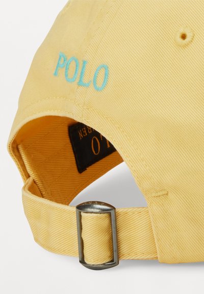 Gul bomullslue med buet skjerm, med "POLO" brodert i lyseblått og en justerbar stoffstropp med en sølvspenne.