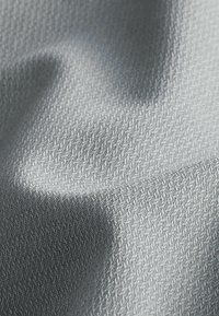 Hellgrauer, strukturierter Stoff mit einem subtilen geometrischen Muster. Glatte Oberfläche mit einem sanften Glanz, der zarte Falten und Schatten erzeugt.