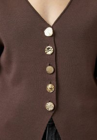Brązowy kardigan z głębokim dekoltem w kształcie V i sześcioma dekoracyjnymi złotymi guzikami, w tym z motywem muszli i teksturowymi akcentami, z bocznymi rozcięciami.