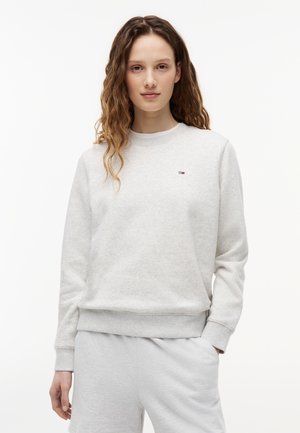 Mladá žena s dlhými kučeravými vlasmi, oblečená v svetlosivej mikine a zodpovedajúcich teplákoch, stojaca pred bielym pozadím.