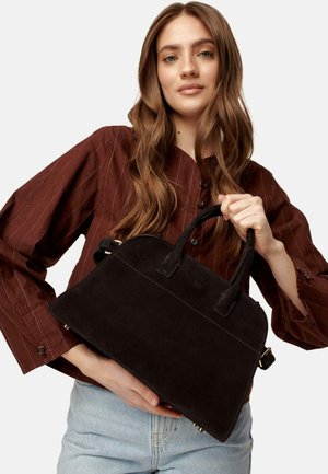 Zwarte suède handtas met gestructureerd ontwerp, dubbele bovenhandvatten en een ritssluiting aan de binnenkant. Model houdt het vast tegen een bruine blouse met knopen.