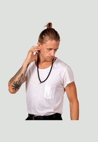 T-shirt bianca a collo rotondo con taschino, collana di perline nere, braccio tatuato, capelli corti raccolti in un chignon e cintura nera.
