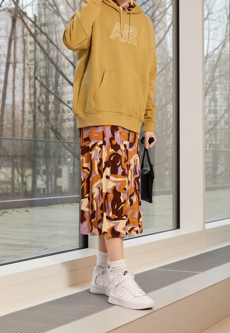 Khaki hooded sweatshirt with front pocket, patterned midi skirt in brown, purple, and orange. White high-top sneakers, holding a black bag.