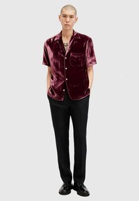 Mężczyzna z łysą głową, ubrany w krótki rękaw bordowej aksamitnej koszuli z guzikami, czarne spodnie i czarne buty eleganckie, stojący z rękami w kieszeniach.