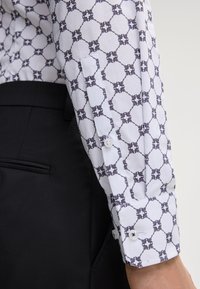 Chemise blanche à manches longues avec un motif géométrique en bleu marine. Présente des poignets à boutons et un tissu texturé. Portée avec un pantalon noir.