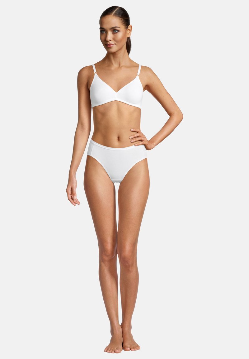 Donna sta in piedi a piedi nudi, indossando un reggiseno bianco e slip a vita alta, posando con una mano sui fianchi davanti a uno sfondo semplice e chiaro.