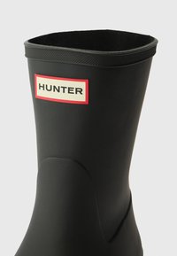 Black rubber knee-high boot with a textured finish; features a red and cream label with the word "HUNTER." Smooth design without visible hardware.