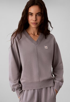 Ung kvinde med langt mørkt hår iført en løs, lyse lilla V-hals sweatshirt og matchende bukser med hænderne i lommerne.