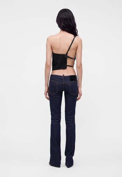 Femme aux longs cheveux foncés, debout de dos, portant un crop top asymétrique noir à bretelles et un jean bootcut bleu foncé.