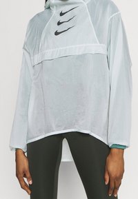 Cazadora ligera azul claro de Nike con capucha, tres logotipos Swoosh negros en el pecho, puños elásticos y un corte relajado.