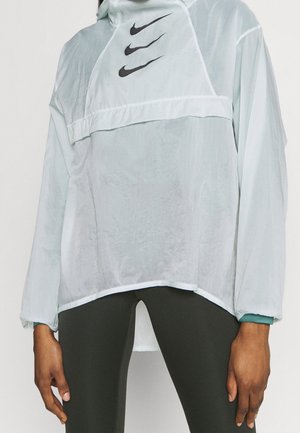 Lysblå Nike vindjakke med hætte, tre sorte swoosh-logoer på brystet, elastiske manchetter og en afslappet silhuet.