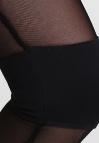Des leggings en maille noire présentent un motif texturé, avec une taille lisse et unie en noir. Le design passe de la maille à un matériau uni.
