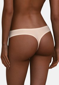 Beige sömlös stringtrosa med en slät textur och kontrasterande midjeband, designad för minimal täckning och komfort.