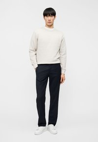 Νεαρός άνδρας στέκεται, φορώντας ανοιχτό μπεζ φούτερ, μαύρο παντελόνι και λευκά αθλητικά παπούτσια, μπροστά σε απλό λευκό φόντο.