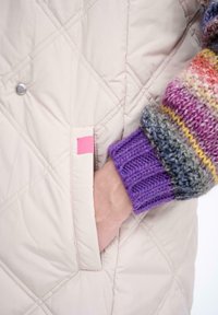 Jasno beżowa pikowana kurtka z boczną kieszenią i różowym akcentem metki, zestawiona z wielokolorowym dzianinowym rękawem z żywymi paskami.