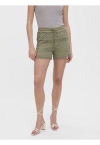 Shorts en dentelle vert olive avec un ourlet festonné et une taille à cordon, associés à des talons nus à brides. Le tissu présente des motifs complexes.